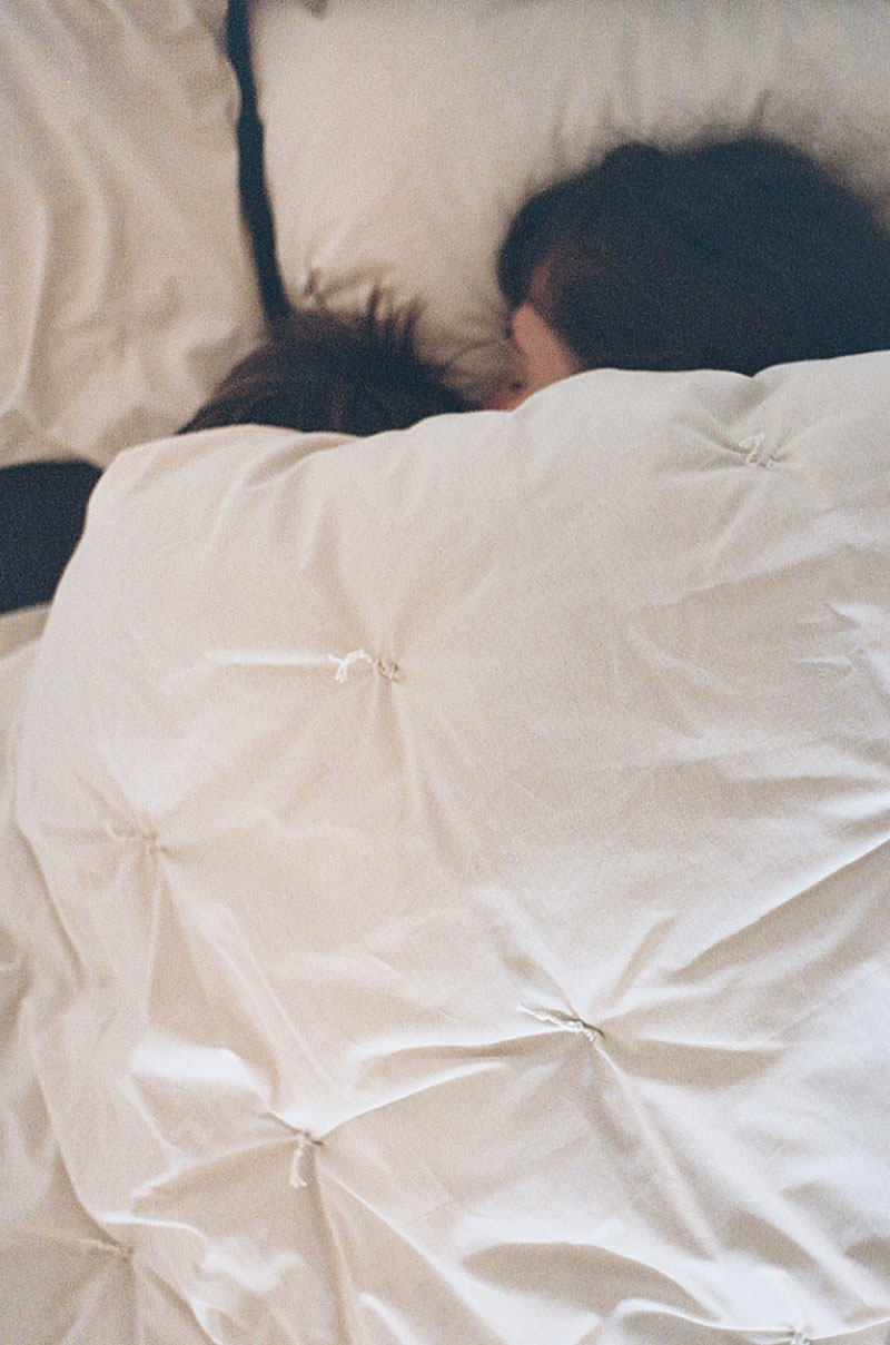 Mutter und Kind liegen schlafend unter einer Bettdecke. Nur das Haar der beiden guckt unter der Decke hervor.