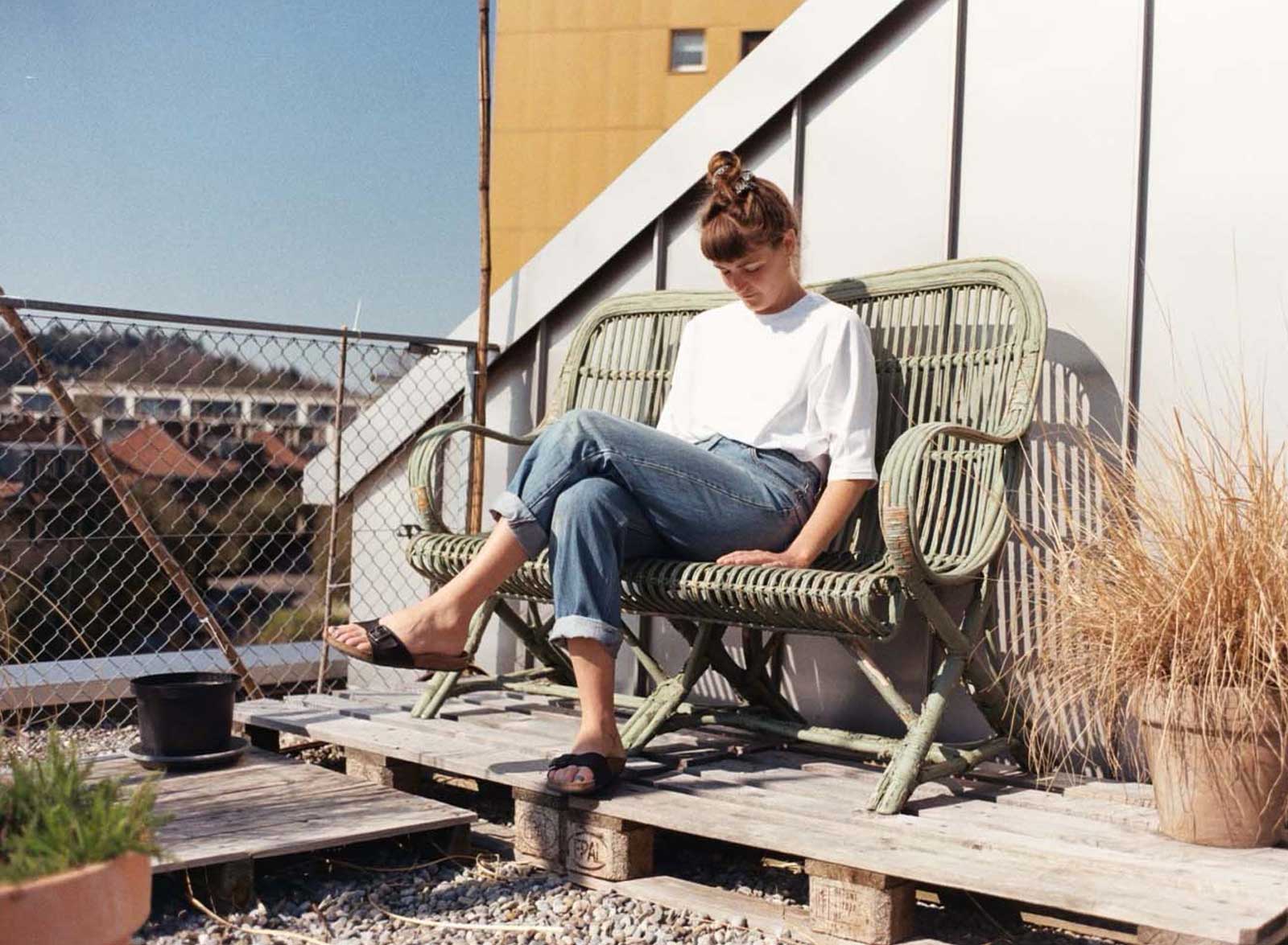 Weibliche Person mit weissem Shirt und Bluejeans sitzt auf einer Sonnenterasse in einer geflechteten Couch.